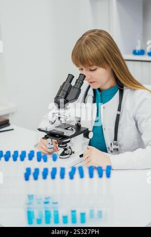 Eine Wissenschaftlerin mit weißem Labormantel arbeitet aufmerksam mit einem Mikroskop in einem Labor. Blaue Reagenzgläser sind im Vordergrund angeordnet Stockfoto