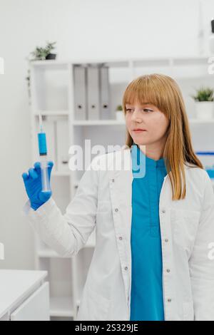 Eine Ärztin in weißem Fell und blauen Handschuhen untersucht sorgfältig eine Spritze mit blauer Flüssigkeit. Der Hintergrund des Labors betont einen Profi Stockfoto