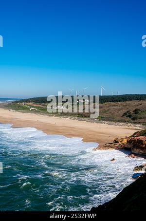 Nazaré, Portugal – stürzende Wellen und Klippen umrahmen Windturbinen auf fernen Hügeln und verbinden natürliche Schönheit mit erneuerbarer Energie. Stockfoto