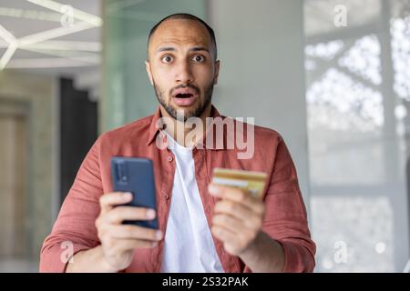 Porträt eines enttäuschten und getäuschten Mannes, der die Kreditkarte und das Telefon in der Hand hält, das Konto blockiert und der Geldtransfer abgelehnt wurde. Betrügerische Handlungen. Stockfoto