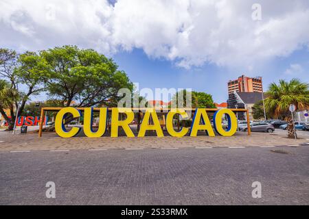 Großes gelbes Curacao-Schild im Freien mit Bäumen, Gebäuden und geparkten Autos im Hintergrund. Willemstad. Curacao. Stockfoto