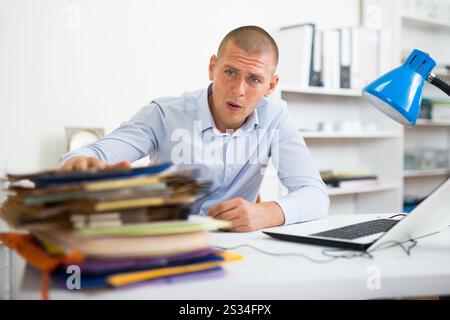 Schockierter Geschäftsmann, der mit vielen Papiermappen am Bürotisch saß Stockfoto