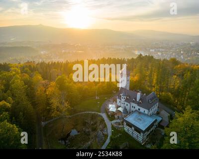 Der Liberecka Vysina Aussichtsturm erhebt sich majestätisch in leuchtenden Herbstfarben. Die Sonne untergeht hinter den Bergen und strahlt ein warmes Leuchten über die malerische Landschaft und die Stadt Liberec. Stockfoto