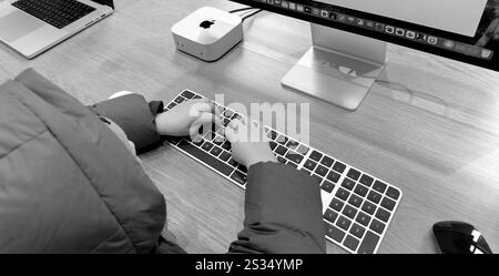 Paris, Frankreich - 10. Dezember 2024: Ein Kind, das auf einer Computertastatur auf einem Holztisch eine Jacke trägt. Ein Computermonitor, Apple Mac Mini und l Stockfoto