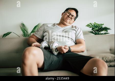 Junger fette Mann in Brille, der auf dem Sofa sitzt, fühlt sich müde, nachdem er Hantel zum Sport benutzt hat, Kopierraum Stockfoto