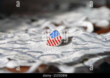 Eine herzförmige amerikanische Flaggennadel sitzt auf einem weißen Tuch. Die Fahnennadel ist aus Silber und hat ein patriotisches Design. Patriotismus und Stolz Stockfoto