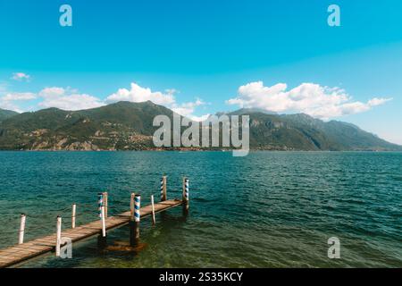 Ruhige Szene am Comer See mit einem verwitterten Holzpier, der sich in ruhiges Wasser erstreckt, mit dem bezaubernden Dorf Menaggio und den üppigen Bergen Stockfoto