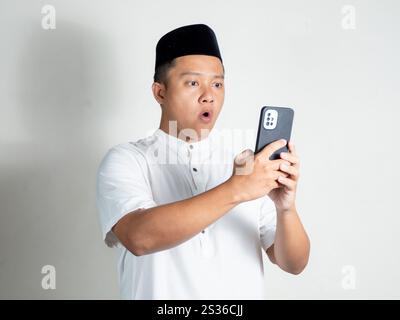 Porträt eines muslimisch-asiatischen Mannes zeigt einen schockierten und überraschten Ausdruck, während er das Telefon hält Stockfoto
