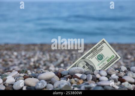 Hundert Dollar, die zur Hälfte mit runden Felsen bedeckt sind, liegen am Strand aus nächster Nähe. Dollarscheine teilweise in Küstensteinen vergraben. Konzept Finanzierung Geld Urlaub Stockfoto