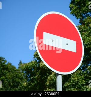 Ein kreisförmiges rotes Schild mit einem weißen Balken, der auf einem grauen Metallpfosten vor grünen Bäumen und blauem, klaren Himmel steht. Ein kreisförmiges rotes Zeichen mit einem Stockfoto