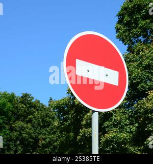 Ein kreisförmiges rotes Schild mit einem weißen Balken, der auf einem grauen Metallpfosten vor grünen Bäumen und blauem, klaren Himmel steht. Ein kreisförmiges rotes Zeichen mit einem Stockfoto