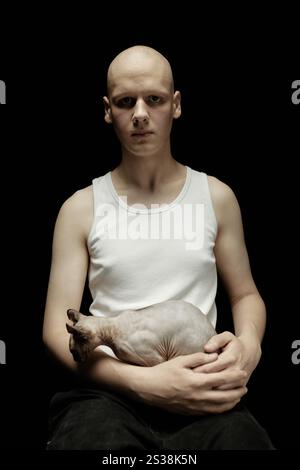 Vertikales Porträt eines jungen Mannes mit hellem Teint, der in weißem Tank-Top gekleidet ist und die haarlose sphynx-Katze auf dem Schoß hält und mit unheimlichem Blick in die Kamera blickt, vor schwarzem Hintergrund im Studio Stockfoto