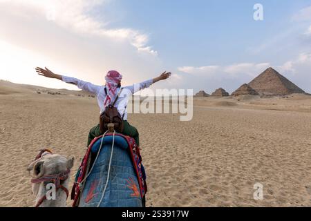 Kamele in der Gizeh Pyramidenanlage - Eine Frau mit einem roten Turban und bewaffneten Armen, die auf einem Kamel durch dünne Sanddünen reiten - Kairo, Ägypten Stockfoto