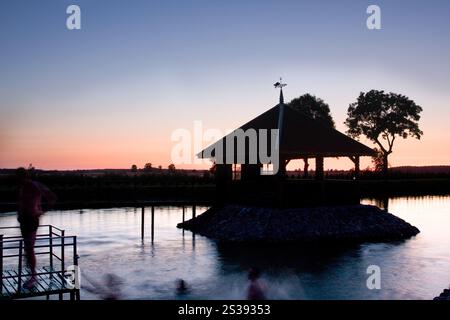 Eine friedliche Seeszene mit Pavillon und schwimmenden Menschen in Silhouette, umgeben von Bäumen und einem ruhigen Sonnenuntergang. Stockfoto