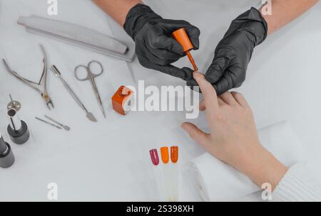 Ein professioneller Manikürer, der lebendigen orangefarbenen nagellack auf die Nägel eines Kunden aufträgt. Werkzeuge und Nailart-Muster werden auf einem weißen Tisch angeordnet, wodurch ein entsteht Stockfoto