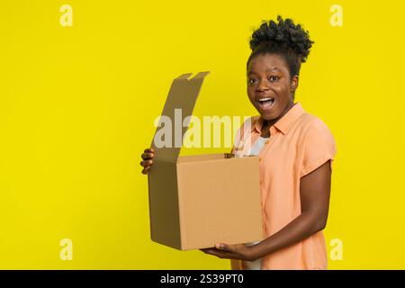 Afroamerikanerin öffnet den Karton und zeigt Aufregung Freude beim Empfang eines Pakets. Schwarzes Mädchen isoliert auf gelbem Hintergrund erhalten schnell d Stockfoto