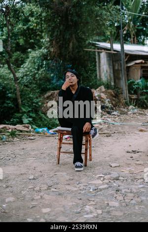 Ein südostasiatischer Mann, der allein auf einem Stuhl sitzt und raucht, sieht schlecht aus Stockfoto