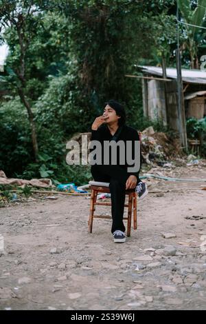 Ein südostasiatischer Mann, der allein auf einem Stuhl sitzt und raucht, sieht schlecht aus Stockfoto