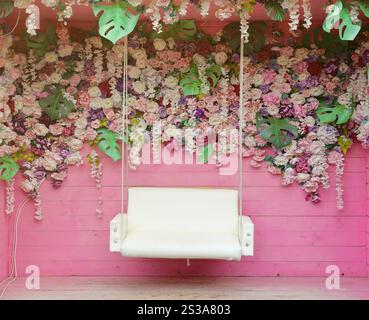 Blumenhintergrund. Viele künstliche Blumen in bunter Komposition auf handgemachtem hölzernem rosa Hintergrund mit weißer Schaukel in der Nähe. Blumenhintergrund. Stockfoto