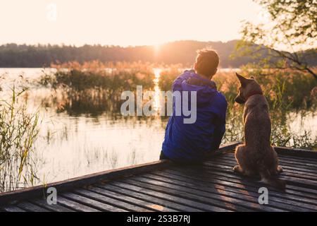 Junger Mann sitzt auf einer Holzterrasse am See und genießt einen wunderschönen Sonnenaufgang mit seinem besten Freund - Ingwerhund Stockfoto