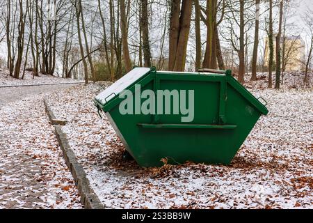 Ein großer grüner Müllcontainer in einem schneebedeckten Stadtpark. Gelbes Laub im Schnee. Stockfoto