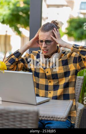 Gutaussehender junger Mann, der Laptop-Computer benutzt, um zu surfen, verliert überraschte plötzliche Lotterieergebnisse, schlechte Nachrichten, Verlust von Vermögen, scheitern. Guy sitzt draußen auf der Terrasse des städtischen Cafés. Lebensstil in der Stadt. Stockfoto