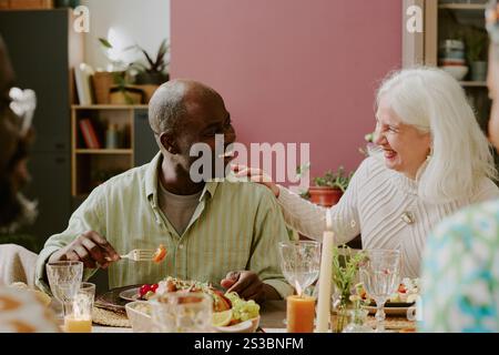 Mittlere Nahaufnahme einer älteren, eleganten Frau, die ihren geliebten afroamerikanischen Ehemann streichelt, während sie essen und sie ansieht Stockfoto