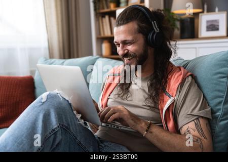 Junger Mann, der von zu Hause aus arbeitet, von zu Hause aus mit einem Laptop arbeitet und Musik über seine kabellosen Kopfhörer hört. Musikexperte und Blogger Stockfoto