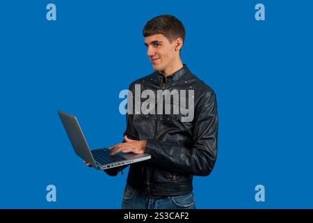 Ein junger Programmierer mit schwarzer Lederjacke benutzt einen Laptop und lächelt, während er auf blauem Hintergrund wegschaut Stockfoto