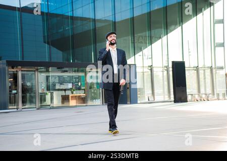 Selbstbewusster Mann, der auf dem Mobiltelefon spricht, während er in der Innenstadt läuft, zeigt Professionalität und lässigen Stil. Junger erwachsener bärtiger Kerl, der ein Handy-Gespräch führt. Männlich mit Smartphone. Hochwertige Fotos Stockfoto