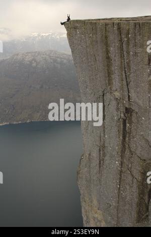 Ein Mädchen sitzt am Rande von Preikestolen in Norwegen, die Hände hoch und überblickt an einem sonnigen Tag den atemberaubenden Fjord. Stockfoto