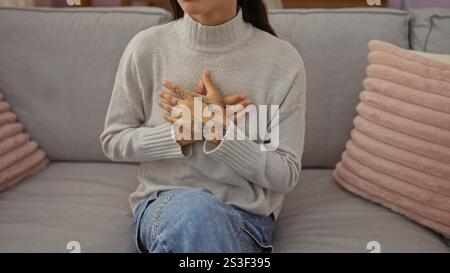 Junge Frau mit Tätowierungen auf den Händen, die im Wohnzimmer des Zuhauses in gemütlicher Inneneinrichtung sitzt, mit Händen auf der Brust in einer nachdenklichen Pose Stockfoto