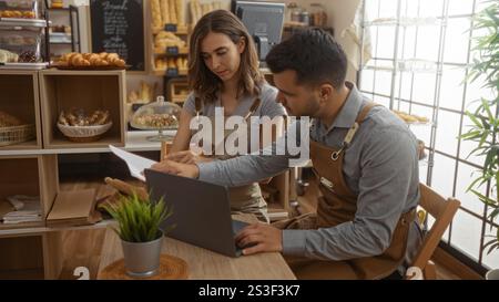 Mann und Frau arbeiten in einer Bäckerei zusammen, tragen Schürzen und benutzen einen Laptop mit Backwaren im Hintergrund, was ein Kollabo anzeigt Stockfoto