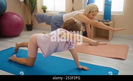 Mutter und Tochter üben gemeinsam Yoga auf Matten in einem hell beleuchteten Fitnessstudio mit Trainingsgeräten im Hintergrund Stockfoto