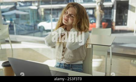 Frau im Büro mit Laptop, die Nackenbeschwerden hat, hebt den Stress am Arbeitsplatz hervor und bietet eine Inneneinrichtung mit sanftem Licht und Kaffee Stockfoto