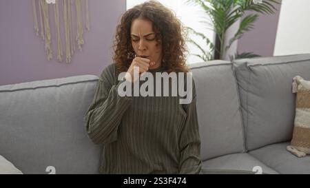 Frau hustet drinnen auf einem bequemen Sofa in einer gemütlichen Wohnzimmerumgebung, drückt Unbehagen mit geschlossenen Augen aus und eine Hand, die ihren Mund bedeckt, surr Stockfoto