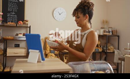 Hispanische Frau, die in einer Bäckerei mit einer Schürze in einem ordentlich organisierten Geschäft mit einem digitalen Tablet auf der Theke arbeitet und Gebäck arrangiert. Stockfoto