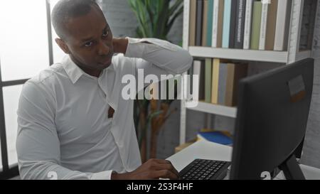 Junger Mann im Büro, der den Hals hält, während er auf den Computer blickt, ein weißes Hemd trägt, drinnen sitzt und Unannehmlichkeiten andeutet, umgeben von Bücherregalen und Stockfoto