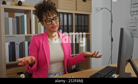 Eine junge Frau in einem rosa Blazer, die an einem Schreibtisch in einem Büro sitzt, mit Regalen und Büchern dahinter Stockfoto