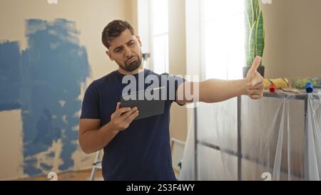 Der junge Mann gibt die Daumen hoch, während er ein Tablet in einem neu bemalten Wohnzimmer benutzt, was auf Zufriedenheit mit dem Fortschritt hinweist, umgeben von Malersup Stockfoto