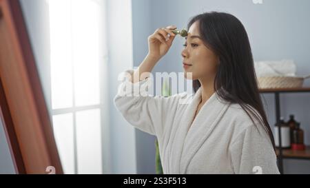 Schöne junge chinesin in einem Spa, die einen Bademantel trägt und einen Jaderoller in einem Wellnesscenter benutzt. Stockfoto