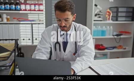 Gutaussehender hispanischer Mann, der an einem Laptop in einem modernen Klinikinnenraum mit medizinischen Geräten arbeitet und eine professionelle und fokussierte Atmosphäre vermittelt. Stockfoto