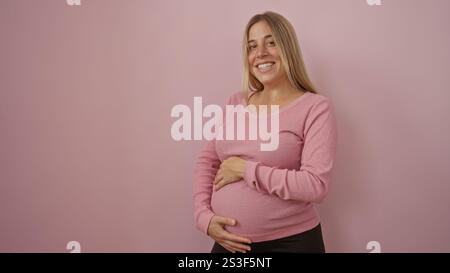 Schwangere Frau mit blonden Haaren in rosa Pullover lächelt über isoliertem rosa Hintergrund und hüllt sanft ihren Bauch und repräsentiert Freude und Erwartung in A Stockfoto