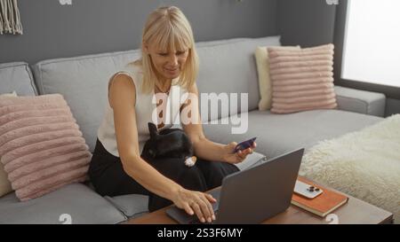 Blonde Frau mittleren Alters mit einem chihuahua auf dem Schoß, der in einem gemütlichen Wohnzimmer einen Laptop benutzt und dabei eine Kreditkarte hält. Stockfoto