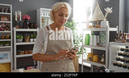 Blonde Frau mit Smartphone im Dekorationsgeschäft mit Regalen mit verschiedenen Produkten im Hintergrund, mit Schürze und im gut beleuchteten Innenbereich Stockfoto