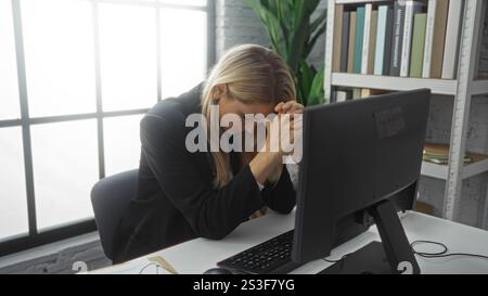 Frau, die am Computer im Büro mit blonden Haaren gestresst ist und drinnen unter natürlichem Licht trauert oder am Schreibtisch mit moderner Inneneinrichtung betet Stockfoto