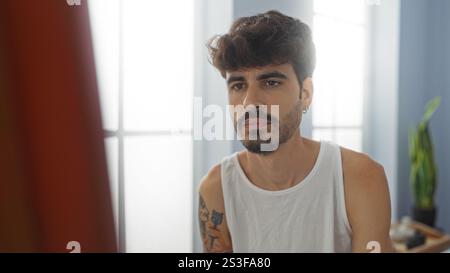 Junger hispanischer Mann mit Bart und Tätowierungen, der drinnen in einem ruhigen Spa-Raum saß und ein weißes Tanktop trug Stockfoto