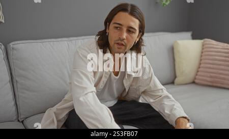 Junger hispanischer Mann mit langen Haaren, der auf einer Couch in einem stilvollen Wohnzimmer sitzt und einen lässigen und doch attraktiven Lebensstil widerspiegelt. Stockfoto