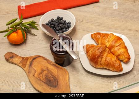 Zwei goldene Croissants sitzen auf einem weißen Teller, neben einem Glas mit dunklem Obstkonfitüre und einer Schüssel Heidelbeeren. Ein Buttermesser und ein hölzernes Schneidebrett Stockfoto