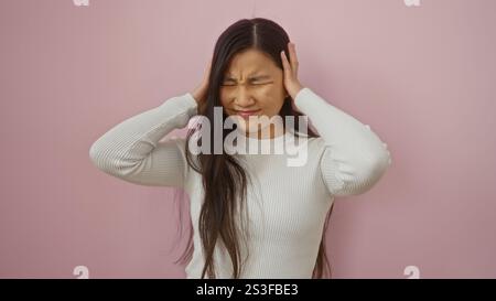 Frau, die Kopfschmerzen empfindet, während sie vor einem sanften rosa Hintergrund steht, Unwohlsein ausdrückt und ihren Kopf berührt. Stockfoto